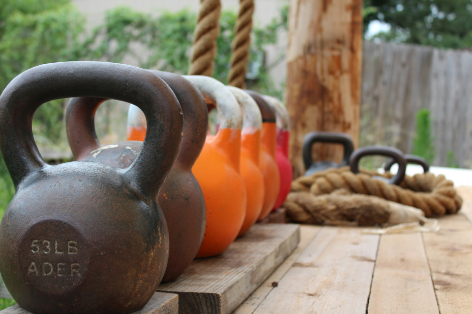 Collection of kettlebells and battle ropes on wooden platform - various weights including 53LB ADER kettlebell, outdoor gym equipment setup